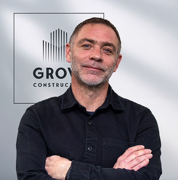 Man with short gray beard, arms crossed, in front of a sign reading 'GROW CONSTRUCTION'.